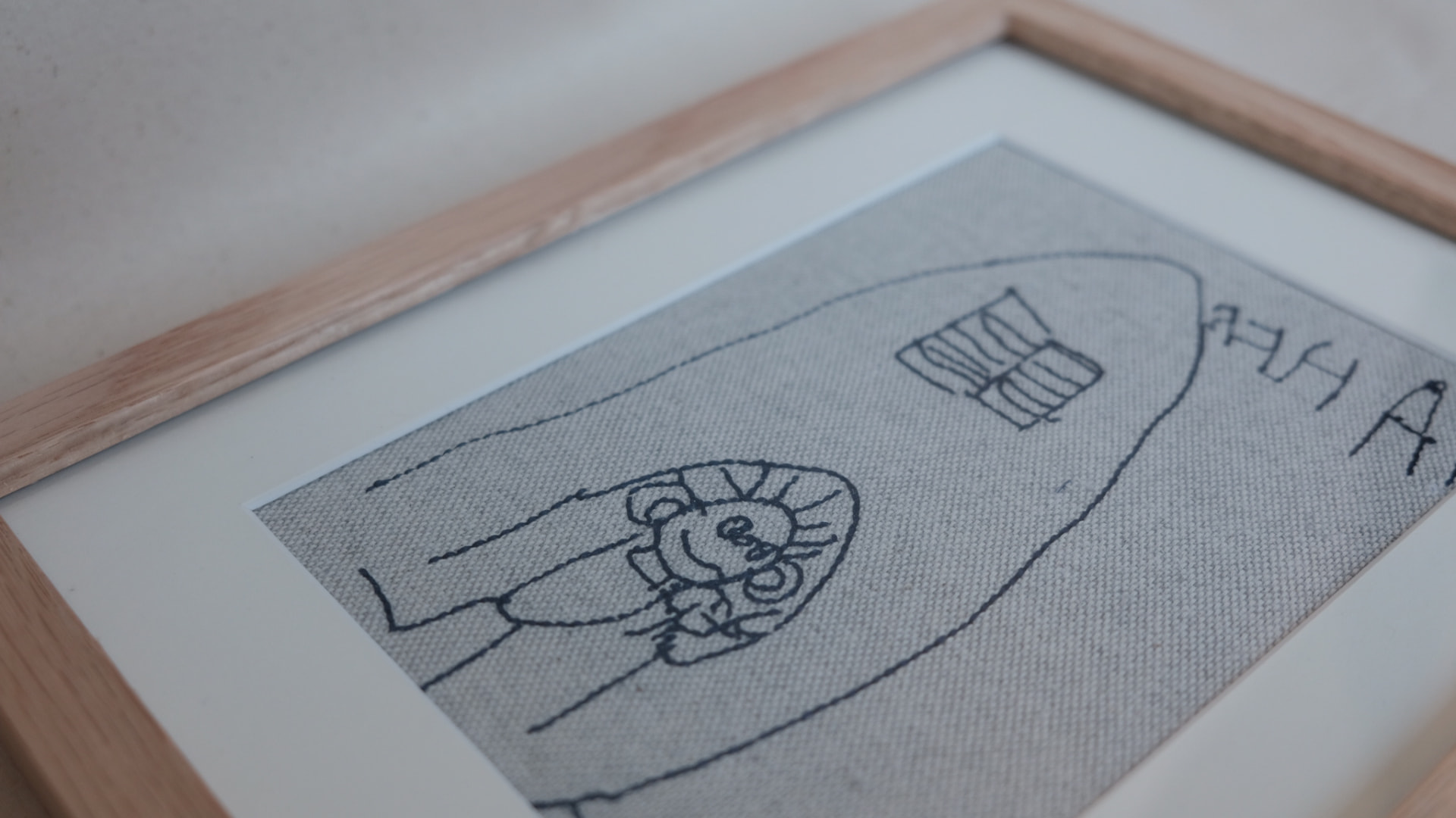 Gestickte Kinderzeichnung mit Haus und Figur, in heller Eiche gerahmt